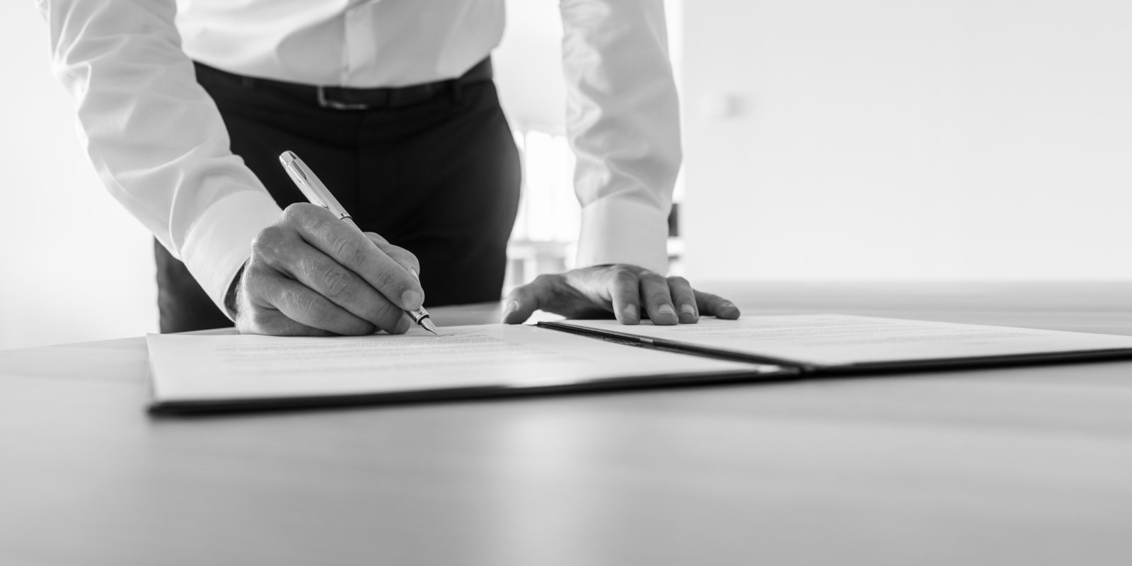 Greyscale image of businessman standing at his desk signing contract or legal document.