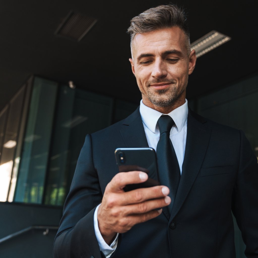 Photo of a positive smiling mature grey-haired handsome business man outdoors at the street near business center using mobile phone.