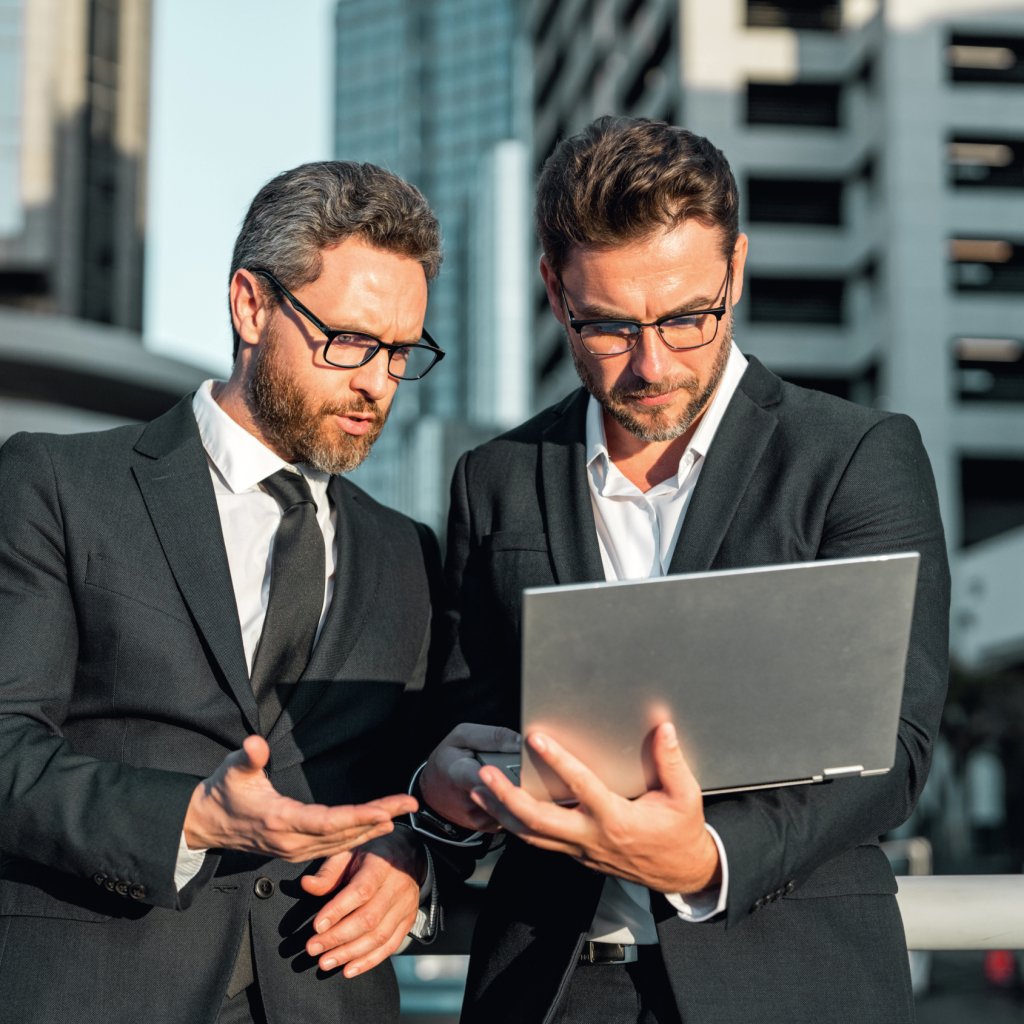 Two handsome young businessmen in classic suits using laptop. Businessmen outdoors near city cityscape using laptop. Businessmen communicating at meeting. Meeting of business partners